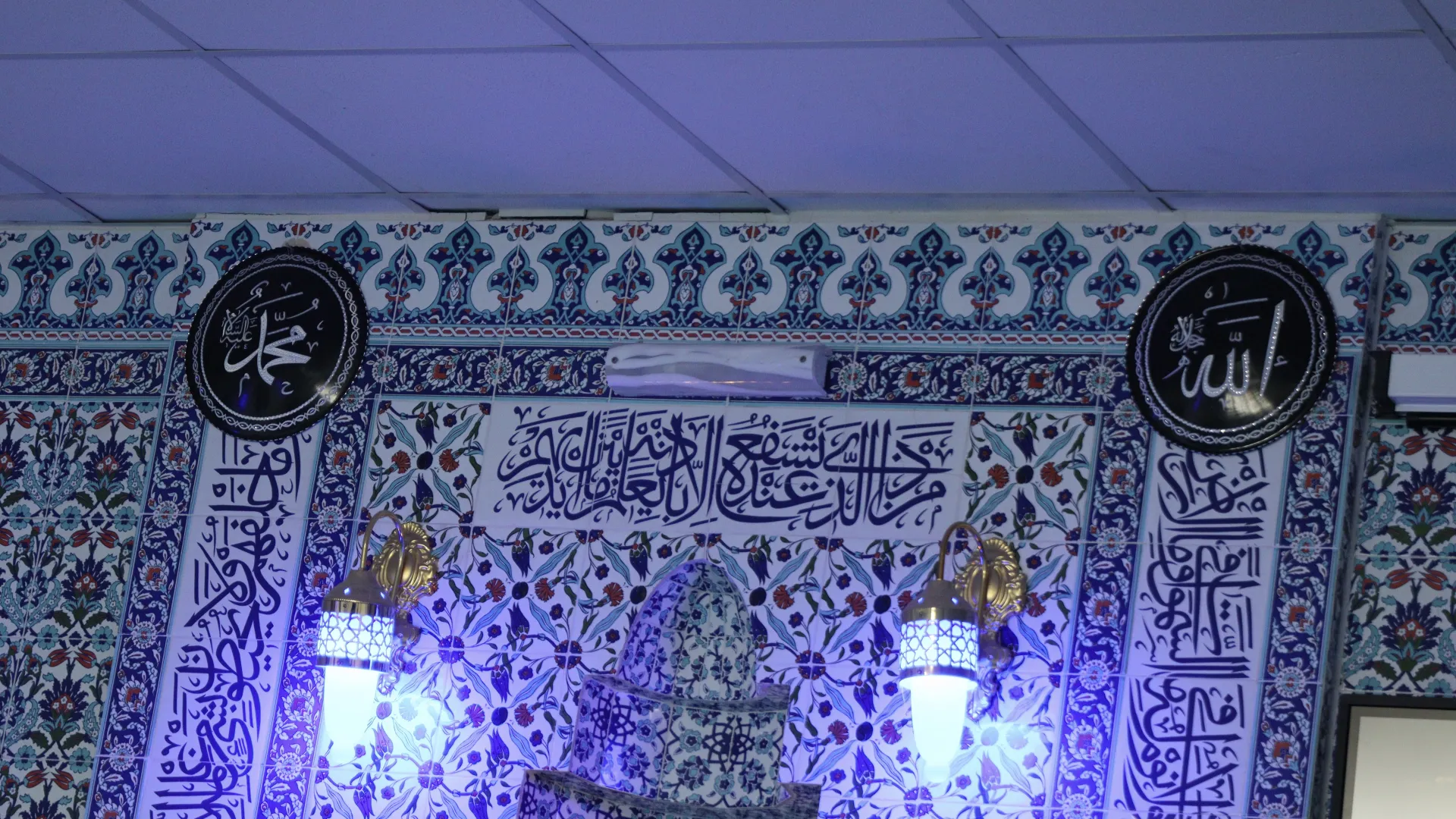 Mihrabın üzerindeki çini duvar: „Muhammed“ ve „Allah“ yazılı yuvarlak hat madalyonları, Arapça kitabe ve altın rengi duvar lambaları