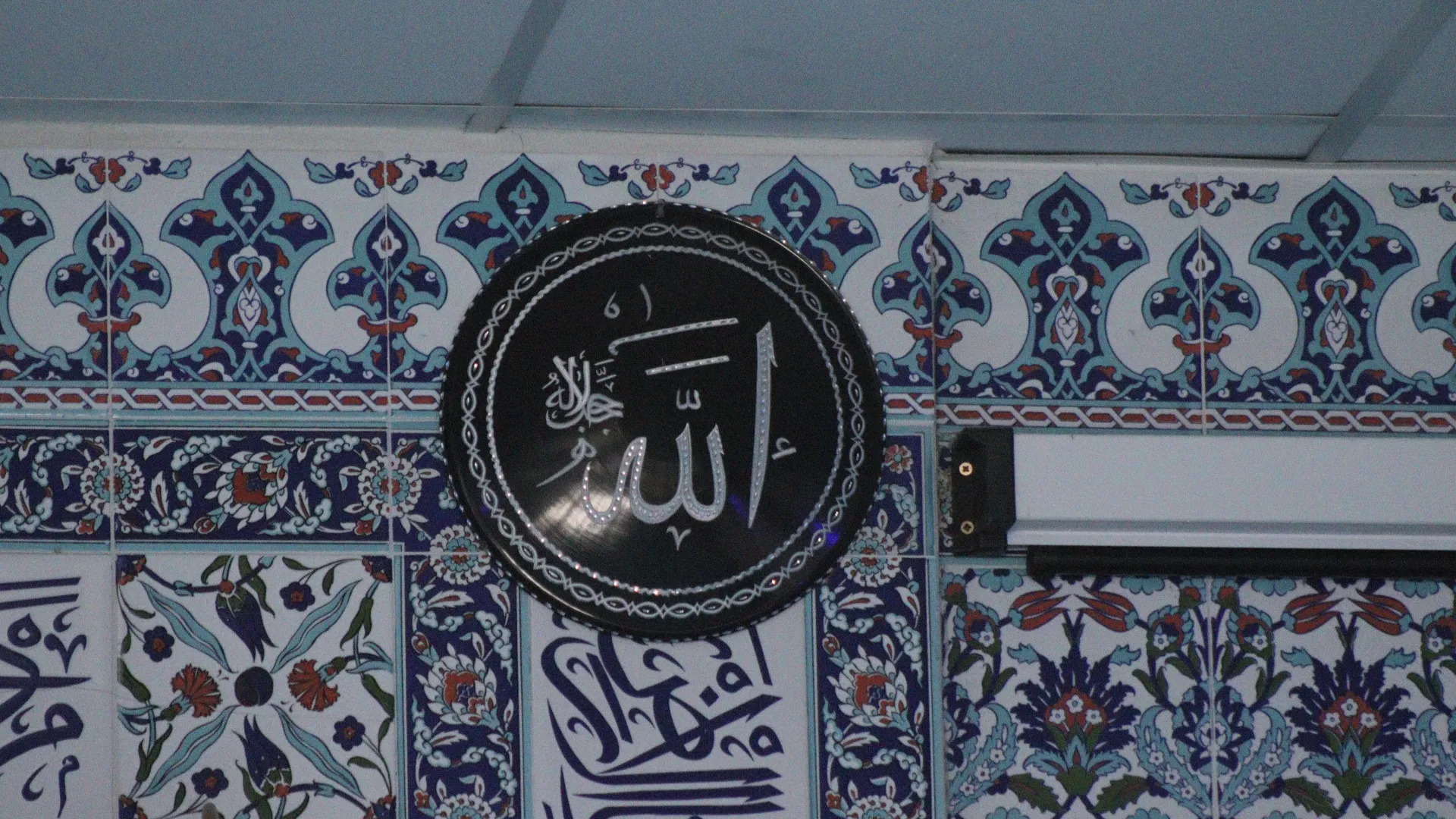 Mavi-beyaz İznik çini duvar önünde „Allah“ yazılı yuvarlak siyah hat madalyonu