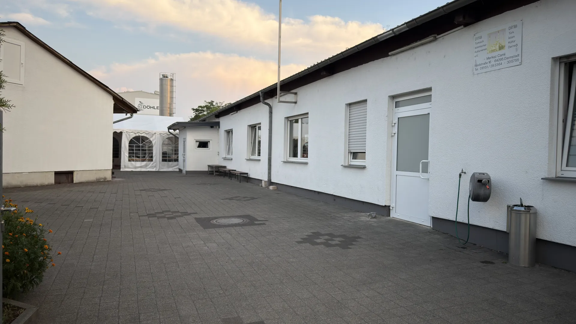 Bina cephesi boyunca uzanan taş döşeli avlu, DITIB dernek tabelası ve oturma bankları