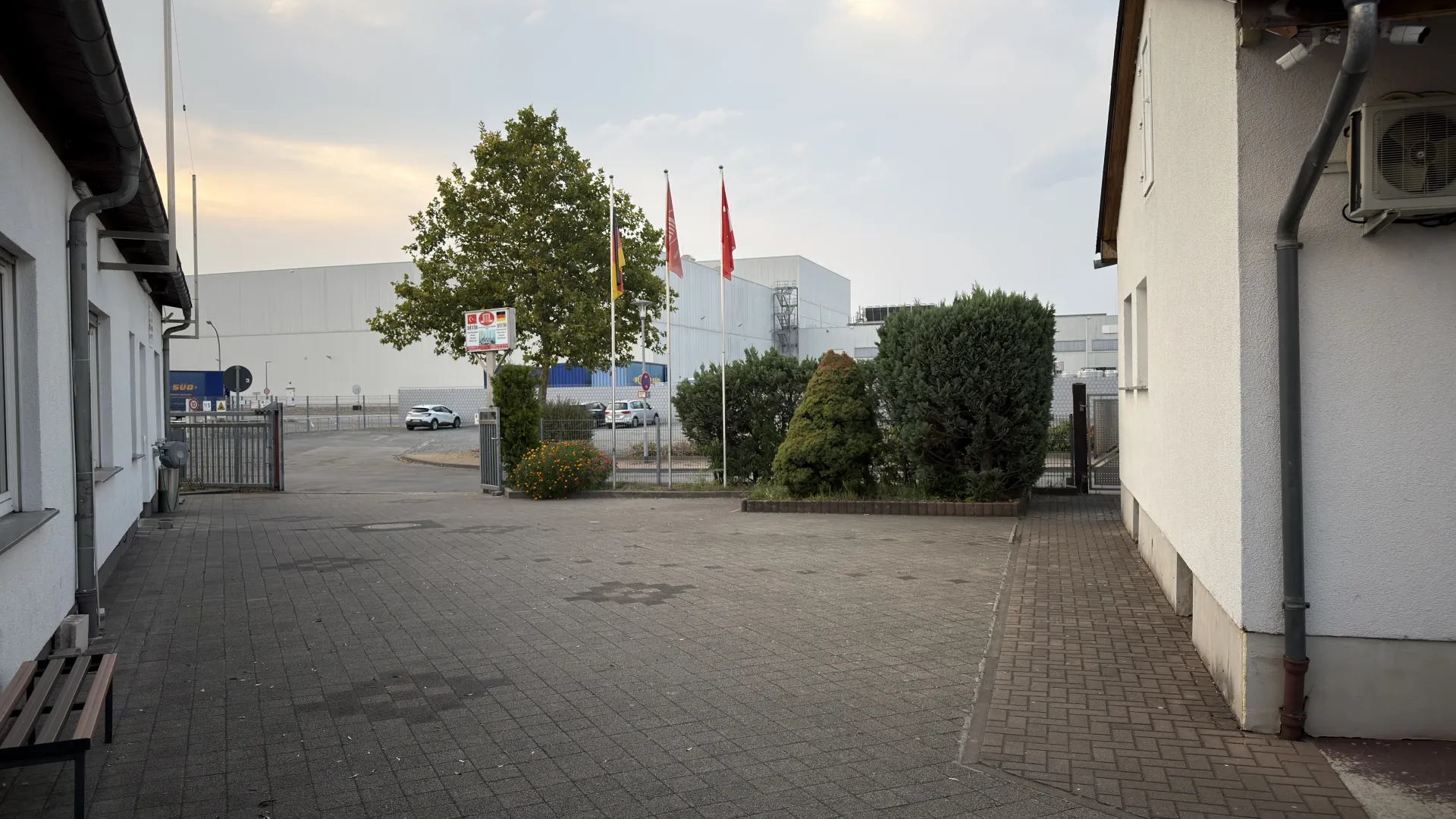 Dernek yerleşkesinin giriş kapısı ve yolu; ağaç, bayraklar ve çiçeklik