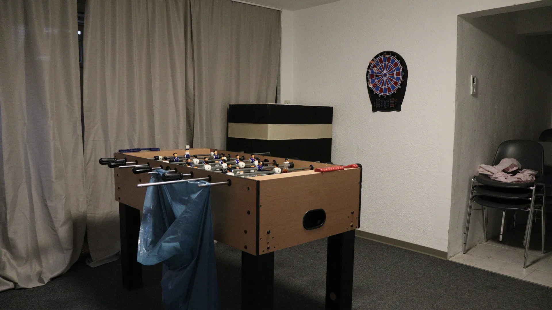 Gençlik odasındaki langırt masası, duvarda elektronik dart tahtası, arka planda istiflenmiş sandalyelerin olduğu geçiş.